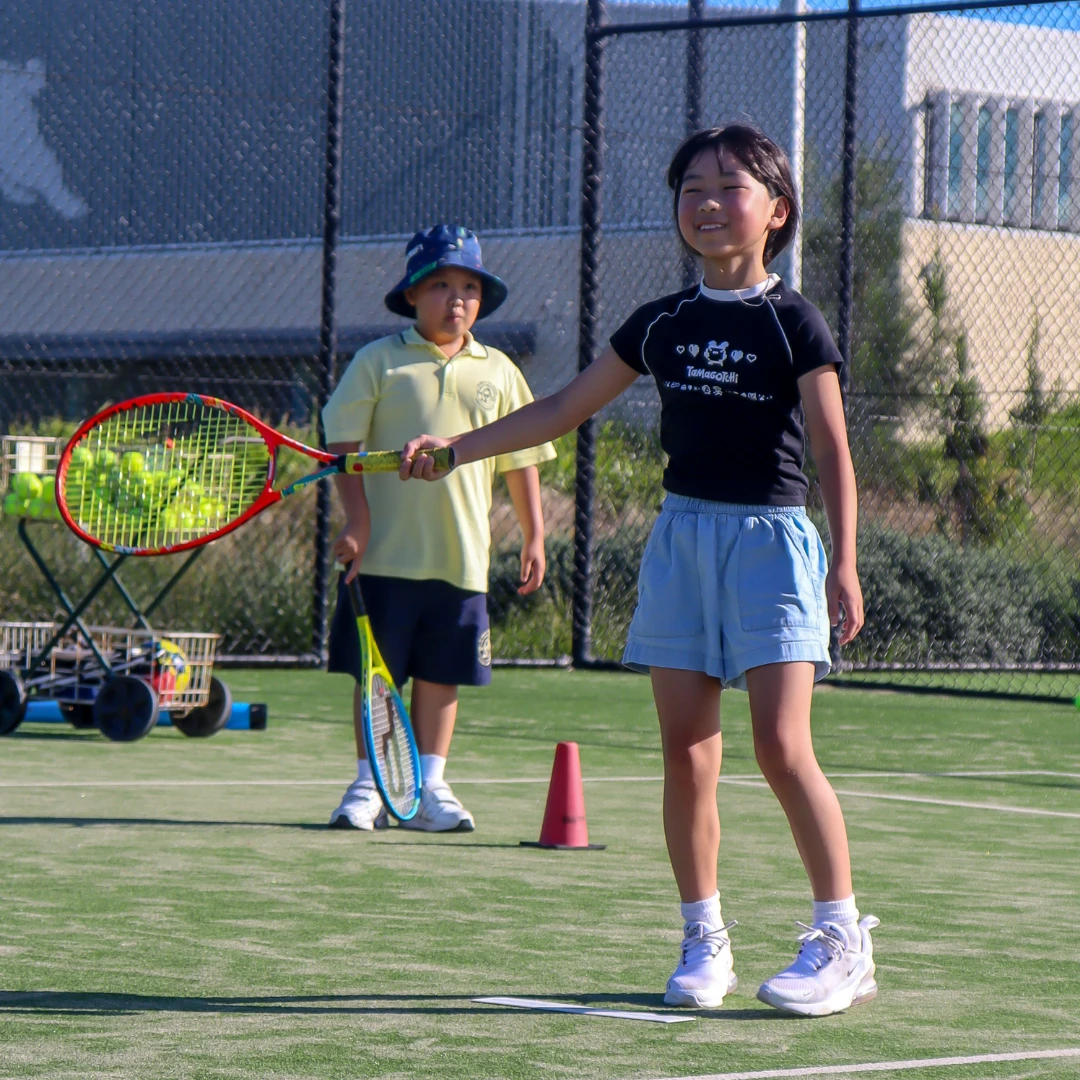 A child standing on an outdoor tennis court holding a tennis racket out in front while smiling.